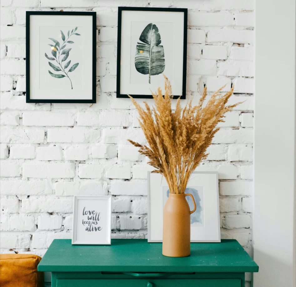 A green side table against a white brick wall, with a small white photo frame reading love will keep us alive standing next to a vase of dried grass