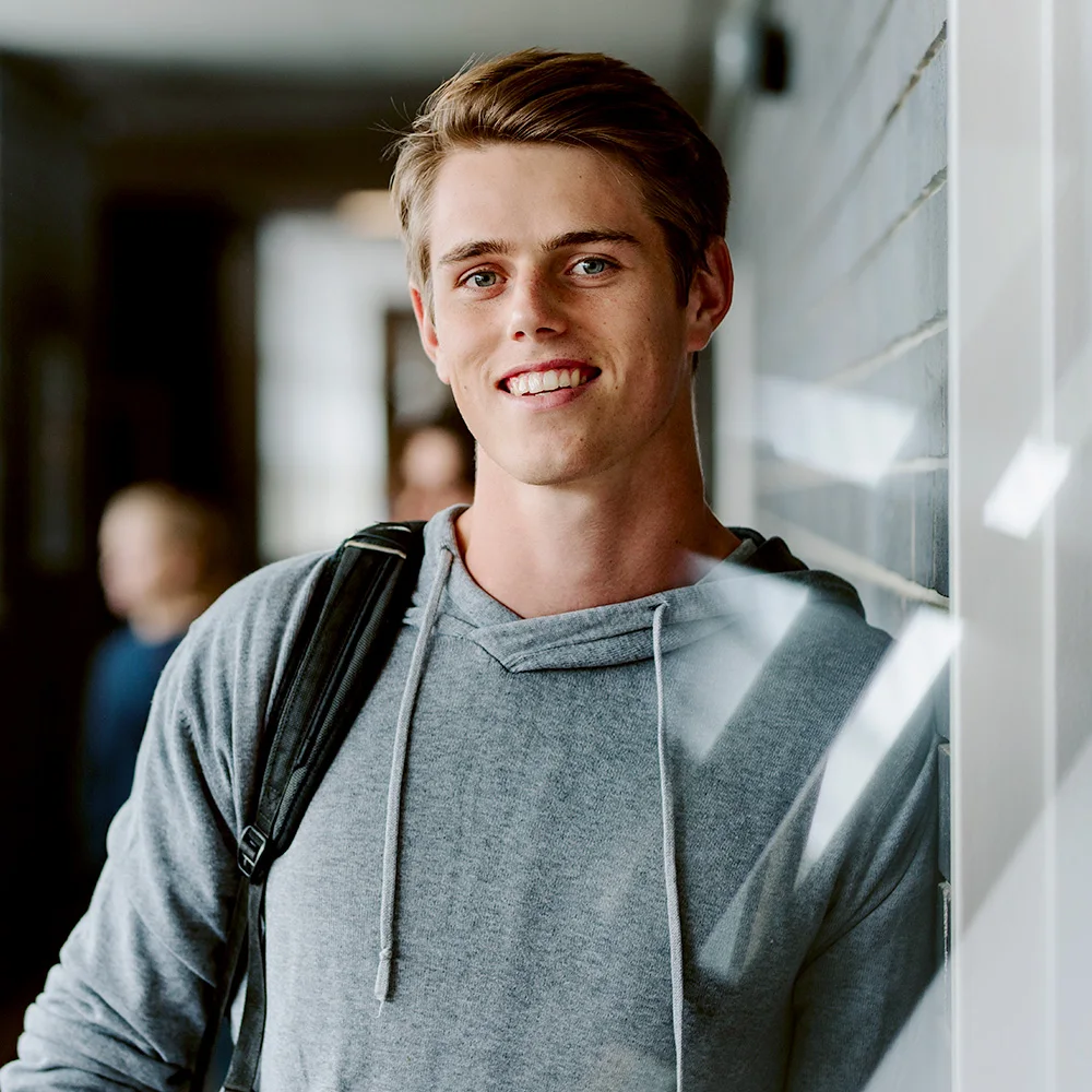 A smiling young man with fair hair in a gray hoodie and backpack, leaning on a hallway wall