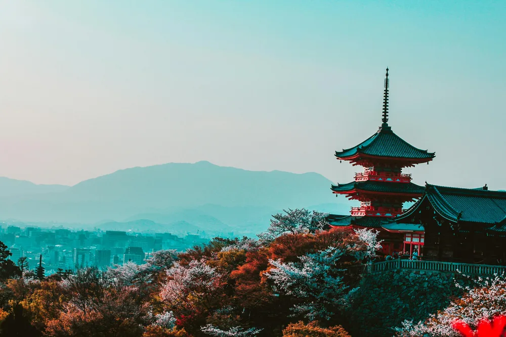 A pagoda on a hillside above a city, with cherry blossom in the foreground and an empty sky above the mountains.