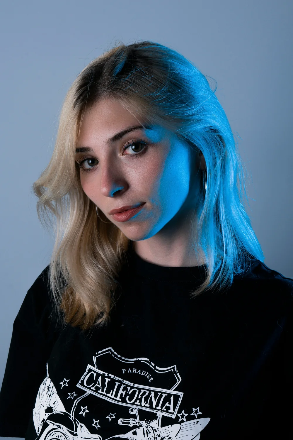 A half-body portrait of a young woman with blond hair in a black T-shirt, lit in blue against a gray backdrop