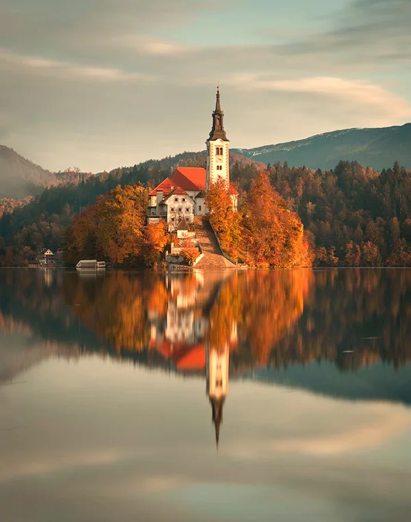 The same lake church with warmer light, a brighter forest and reflection, and a clearer sky