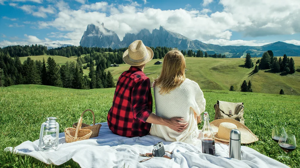 A couple on a picnic blanket in a green meadow, facing mountains under a light blue sky