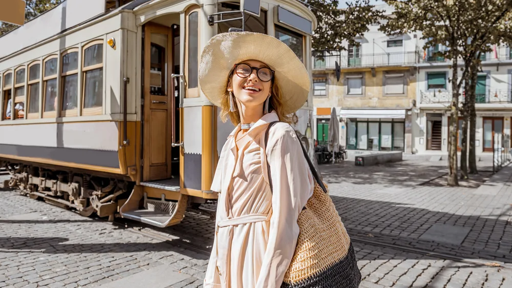 A woman in a straw hat and glasses smiles on a cobbled street with an old tram behind her