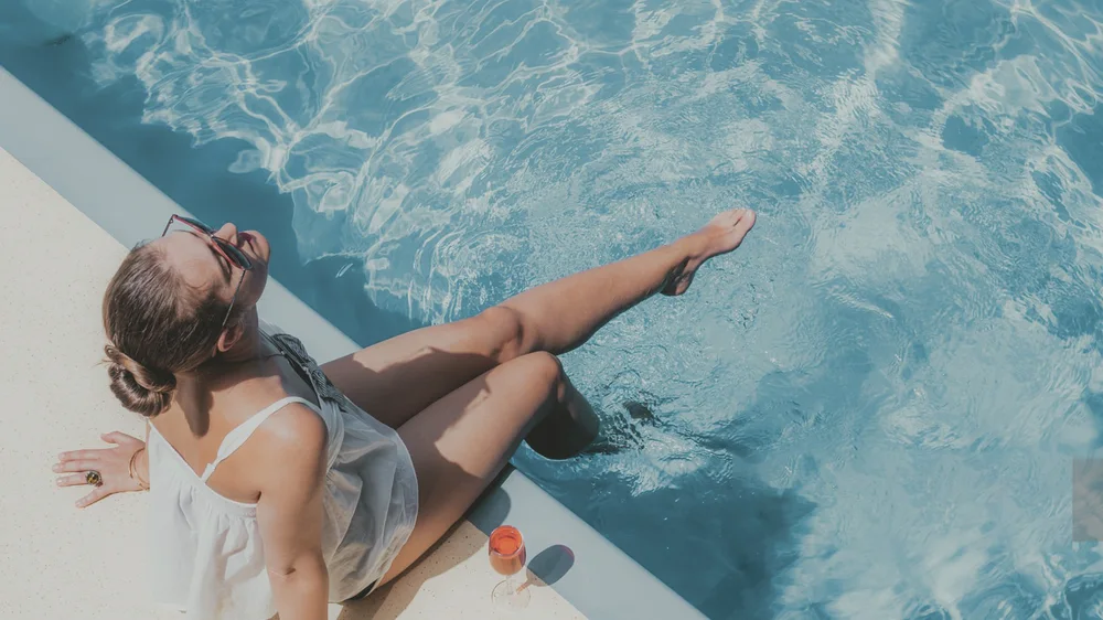A woman in sunglasses sits at the edge of a pool with her feet in the water, in flat, pale colors