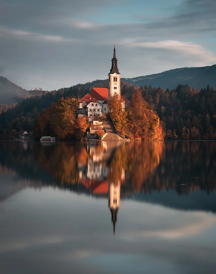 A church on a small lake island with orange autumn trees, reflected in calm water under a gray-blue sky