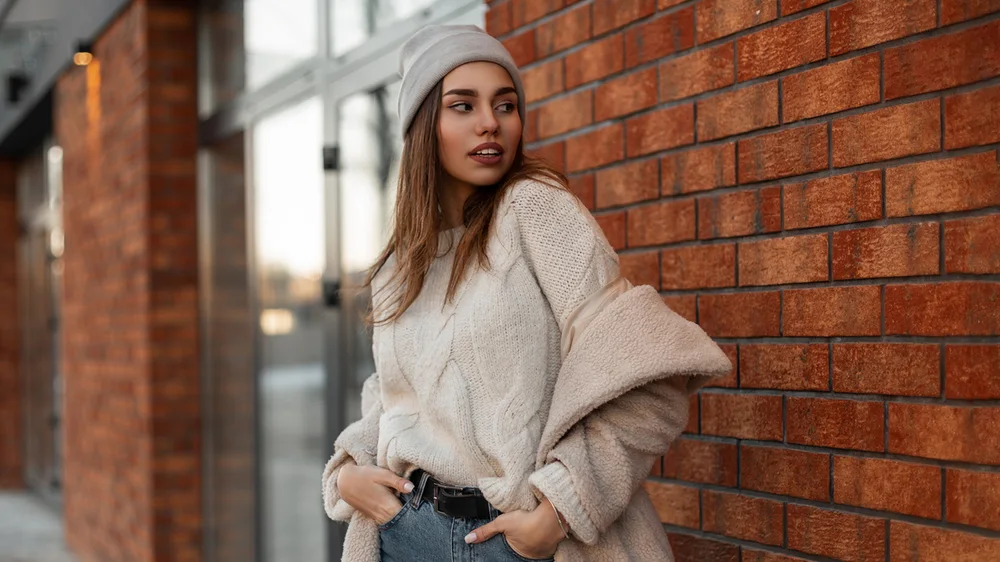 A wide photo of a woman in a knit sweater and beanie against a brick wall, 1920 x 1080 pixels