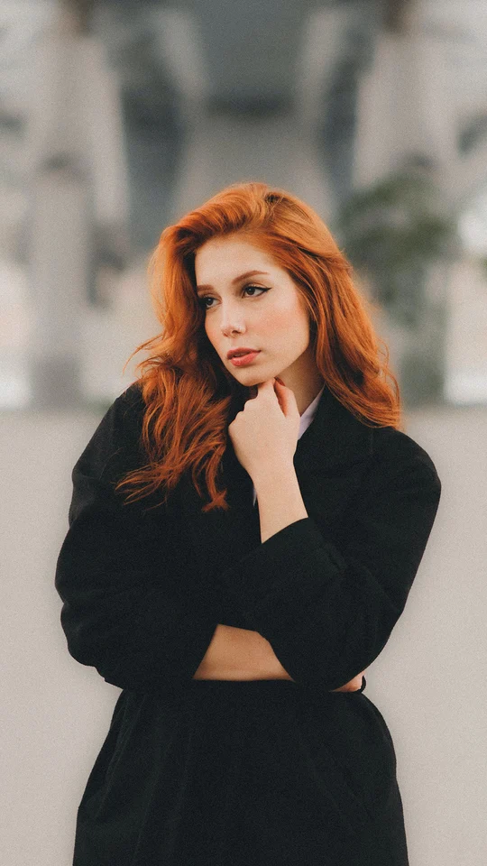 A red-haired woman in a black coat in front of a softly blurred background