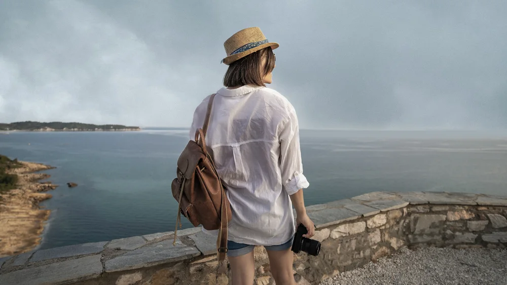 A woman in a straw hat with a backpack looks out over the sea from a stone wall under a flat, overcast sky